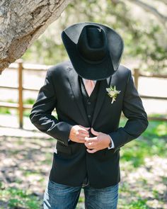 a man wearing a black cowboy hat standing next to a tree in the grass with his hands on his hips