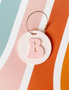 a ceramic ornament with the letter b hanging from it's side on an orange, pink, and blue striped background