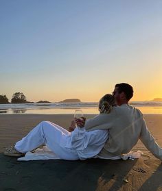 a man and woman sitting on the beach at sunset with wine glasses in their hands