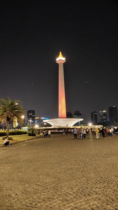 people are standing in the sand at night near a tall tower with lights on it