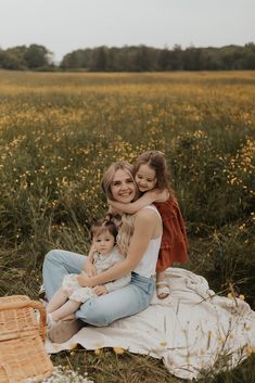 two women and a baby sitting on a blanket in the middle of a field with yellow flowers