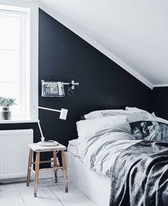 a bed sitting under a window next to a wooden stool in a room with black walls