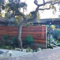 a wooden fence in front of a house with trees and rocks on the side walk