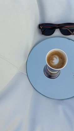 a cup of coffee sitting on top of a blue plate next to sunglasses and a white cloth