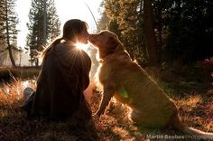 a woman and her dog sitting in the grass with the sun shining through trees behind them