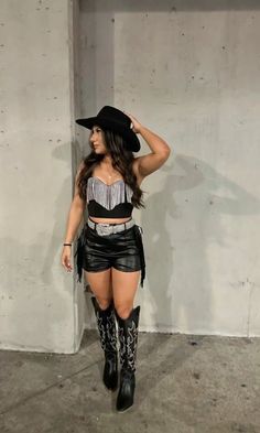 a woman in black leather shorts and cowboy hat standing next to a concrete wall with her hand on her hip