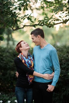 a man and woman standing next to each other in front of some trees with their arms around each other