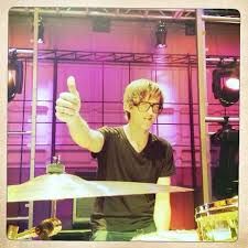 a young man playing drums in front of a purple light and some lights behind him