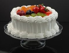 a white cake topped with lots of fruit on top of a glass plate next to a black table