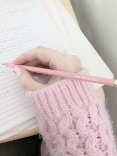 a person holding a pencil in their hand and writing on a notebook with a pink knitted mitt