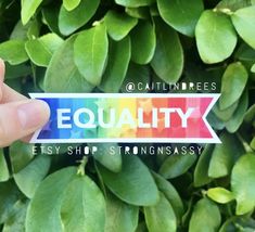 a person holding up a sticker that says equality in front of some green leaves