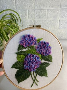 a person holding up a cross - stitch embroidery with purple flowers on it and green leaves