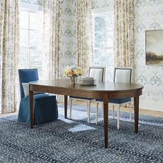 a dining room table with blue chairs in front of floral wallpaper and window curtains