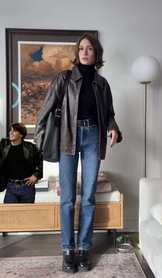 a woman standing in front of a couch wearing jeans and a leather jacket with her hands in her pockets