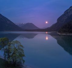 the moon is setting over a lake with mountains in the background