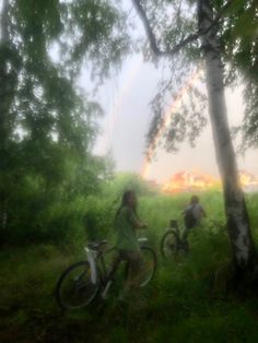 two people riding bikes through the woods with a rainbow in the background