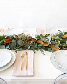 the table is set with white plates and greenery on it, along with silverware