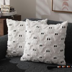 two white pillows with black eyes on them sitting on a gray couch in a living room