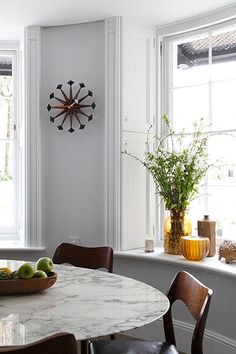 a table with fruit on it in front of two windows and a clock mounted to the wall