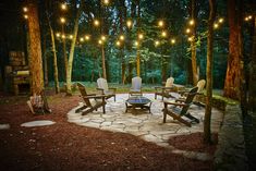 an outdoor fire pit surrounded by chairs and lights in the middle of a wooded area