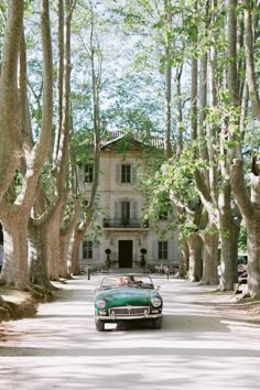 an old green car is parked in front of a large house with trees lining the street