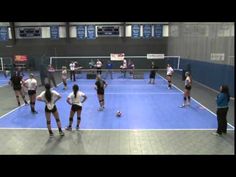 several women are playing tennis on an indoor court with blue and gray flooring, while one woman is holding a racket in her hand