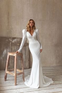 a woman standing next to a stool wearing a white dress with long sleeves and an open back