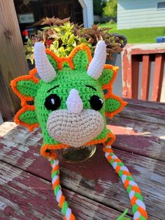 a crocheted green and orange dragon head on a wooden table with plants in the background