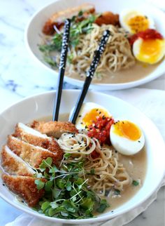 two white bowls filled with noodles, meat and vegetables next to an egg on top