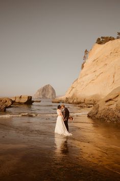 a bride and groom are standing in the water at the beach near some rocks on their wedding day