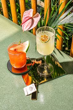 two drinks sitting on top of a green table next to a pink flower and leafy plant