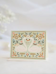 a card with two swans on it and flowers around the edges, in front of a white background