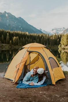 a man and woman sitting in a tent next to each other with mountains in the background