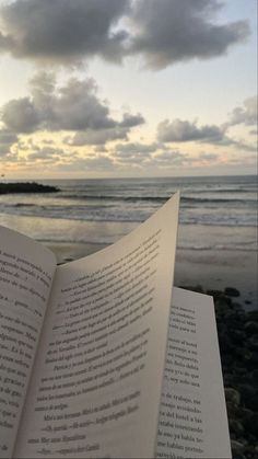 an open book sitting on top of a beach next to the ocean with clouds in the sky