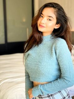 a young woman standing in front of a bed wearing a blue sweater and plaid skirt