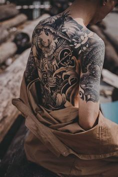 a man with tattoos on his back sitting in front of a pile of wood and looking off into the distance