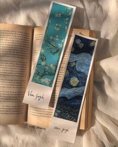 two bookmarks with the same painting on them are laying next to an open book