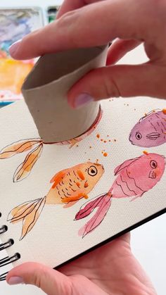 a hand is holding an open notebook with watercolor fish on it