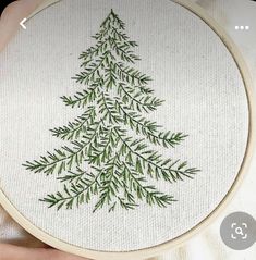 a hand embroidered christmas tree with green leaves on white fabric in front of a person's hand