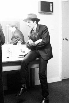 a man in a suit and hat is sitting on a stool looking at himself in the mirror