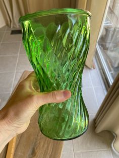 a green glass vase being held up by a person's hand in front of a window