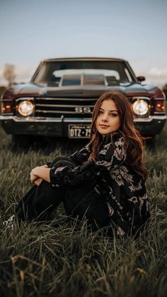 a woman sitting in the grass next to an old car and looking at the camera