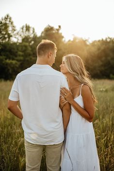 a man and woman standing in tall grass looking into each other's eyes as the sun sets behind them