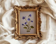 a painting with blue flowers in a gold frame on a white cloth covered bed sheet