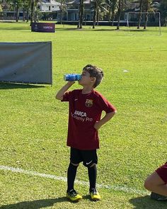 a young boy drinking water from a bottle while standing on a soccer field next to another player