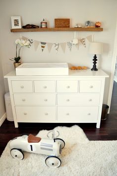 a baby's room with a white dresser and wooden toy car on the floor