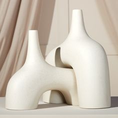 two white vases sitting next to each other on top of a table with curtains in the background