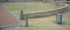 an empty park with benches and a trash can in the foreground, next to a curved brick wall