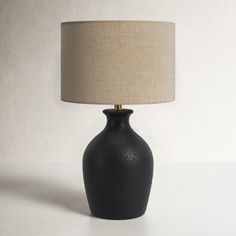 a black table lamp with a beige shade on the base and a white wall behind it