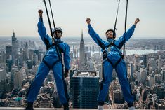 two men in blue jumpsuits standing on top of a building with their hands up